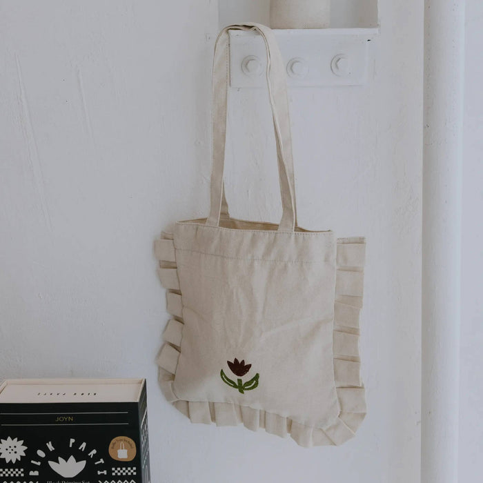 A cream-colored canvas tote bag with ruffled edges and a small flower on the front, hanging on a white wall hook.