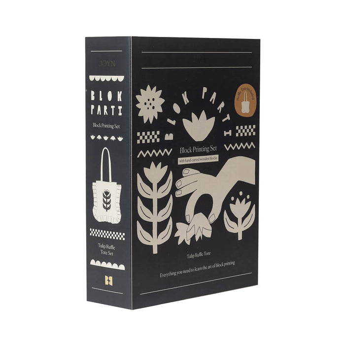 Front angle view of JOYN Blok Parti Tulip Ruffle Tote Block Printing Set, featuring a dark charcoal gray background with cream-colored block printing illustrations of hands, tulip flowers and stem patterns, on a plain white background.