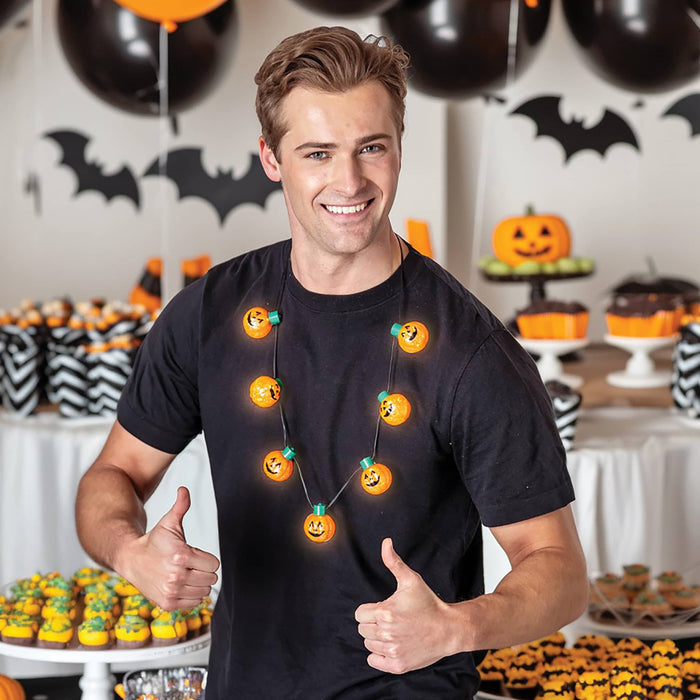 A smiling man wearing a black t-shirt and a glowing orange pumpkin-shaped string light necklace, standing in a decorated Halloween party room with cupcakes, pumpkins, and bat cutouts.