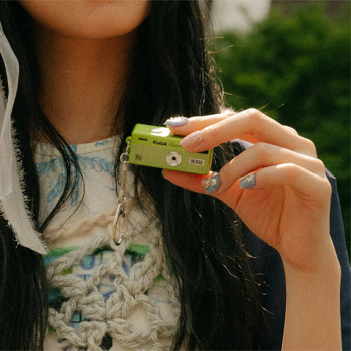 A person holding a small green Kodak Charmera mini digital camera keychain in their hand, with a crochet top and dark hair visible in an outdoor setting.