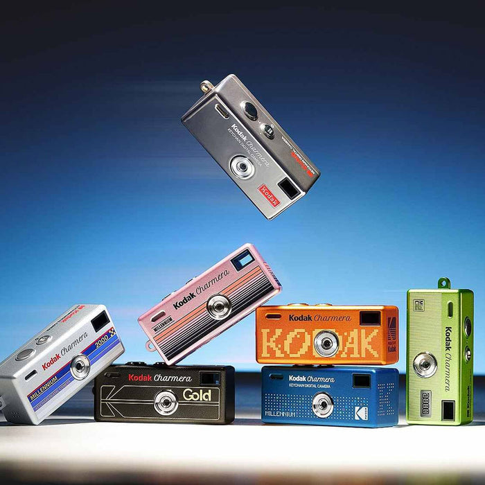 A group of Kodak Charmera mini digital cameras in multiple colors, including silver, pink, orange, black, blue, and green, arranged dynamically against a blue gradient background.