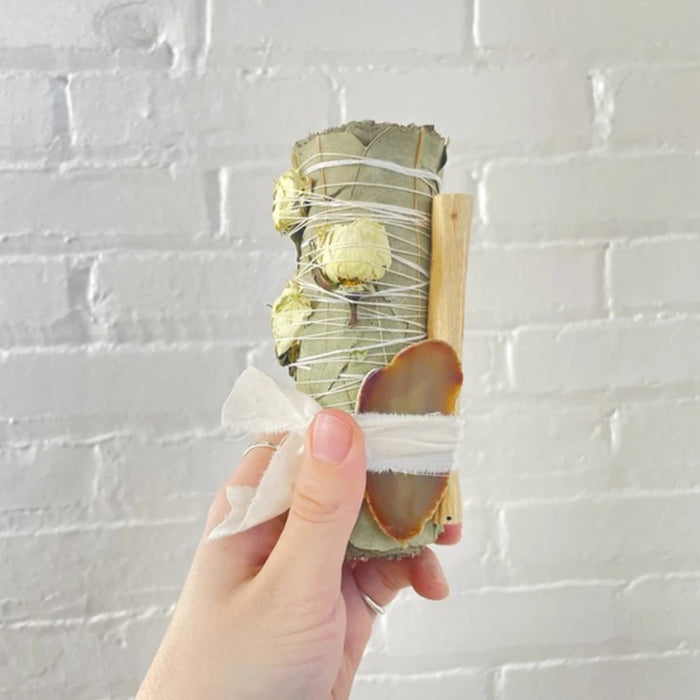 Hand holding a smudge bundle with dried white roses, palo santo and an agate crystal, tied with white fabric ribbon against a white brick wall.