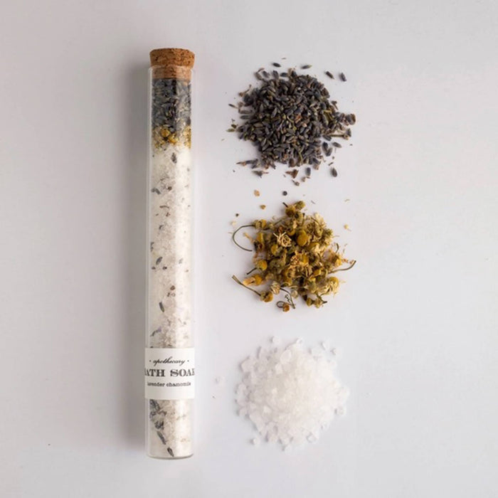 Glass tube with cork stopper filled with bath salt soak with piles of ingredients beside it, on a white background.