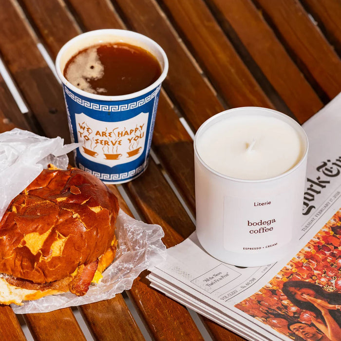 Cup of coffee, a bacon egg and cheese sandwich, and a candle on a wooden table with newspaper.