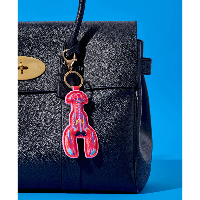 A close-up of a black pebbled leather handbag with a gold-tone oval plate and a lobster-shaped keychain charm in bright pink, red, and blue, set against a solid blue background.