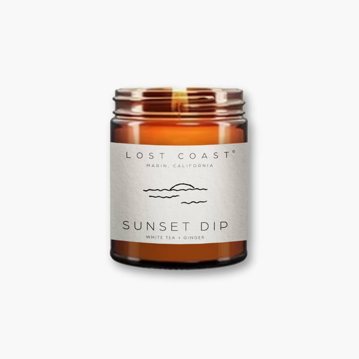 Lost Coast Sunset Dip white tea and ginger scented soy wax blend candle in amber jar, lid off and wick lit, on white background.
