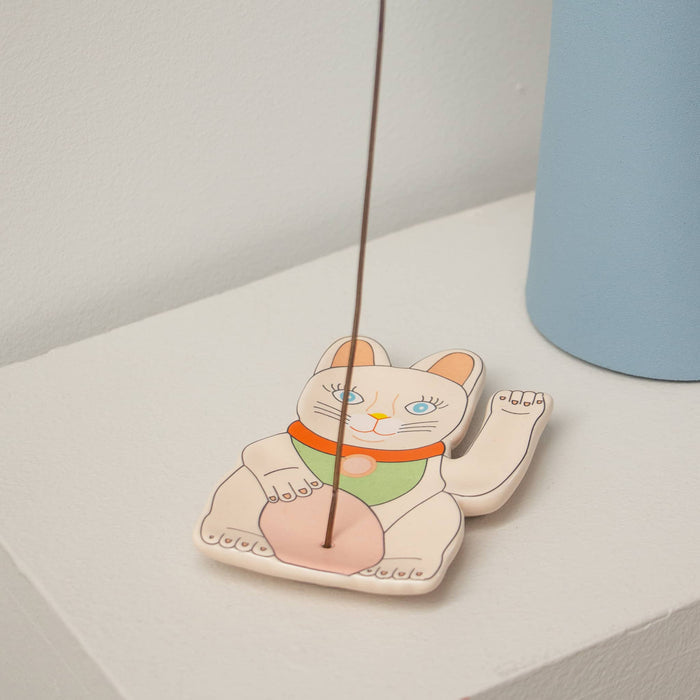 Ceramic incense holder shaped like a lucky cat with an incense stick on a white surface.
