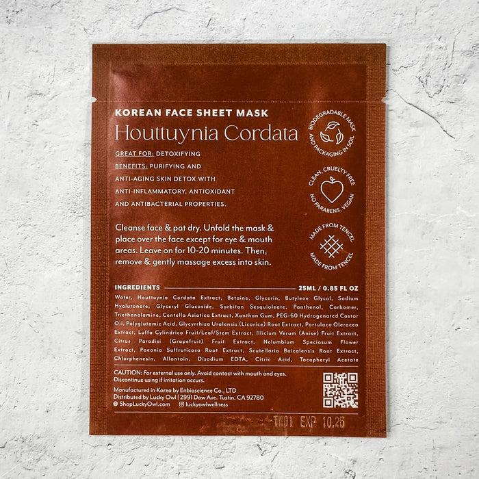 A single brown Korean face sheet mask packet labeled 'Houttuynia Cordata' lies on a light textured white surface. The packet contains product information, ingredients, and a QR code, on a textured backdrop.