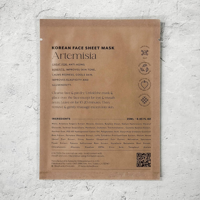 A brown pouch labeled 'Korean Face Sheet Mask Artemisia' with product text, icons, ingredients, and a QR code, photographed against a white textured wall.