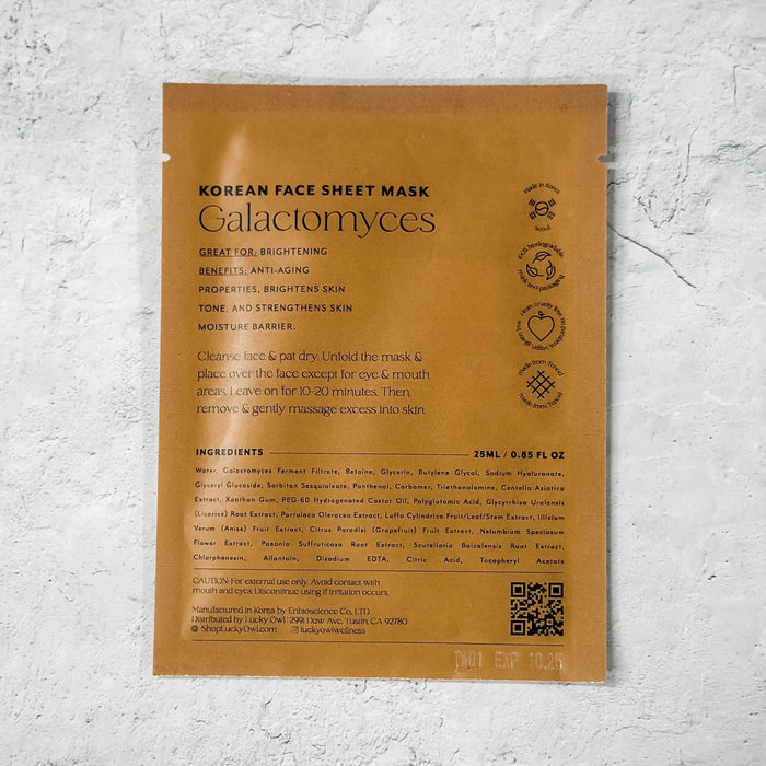 A single-use Korean face sheet mask packet labeled 'Galactomyces' lies flat on a textured white wall. The packet is a warm caramel color with black text detailing the product's benefits, ingredients, and usage instructions.