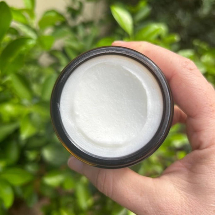 Hand holding a jar of white cream against a green leafy background.