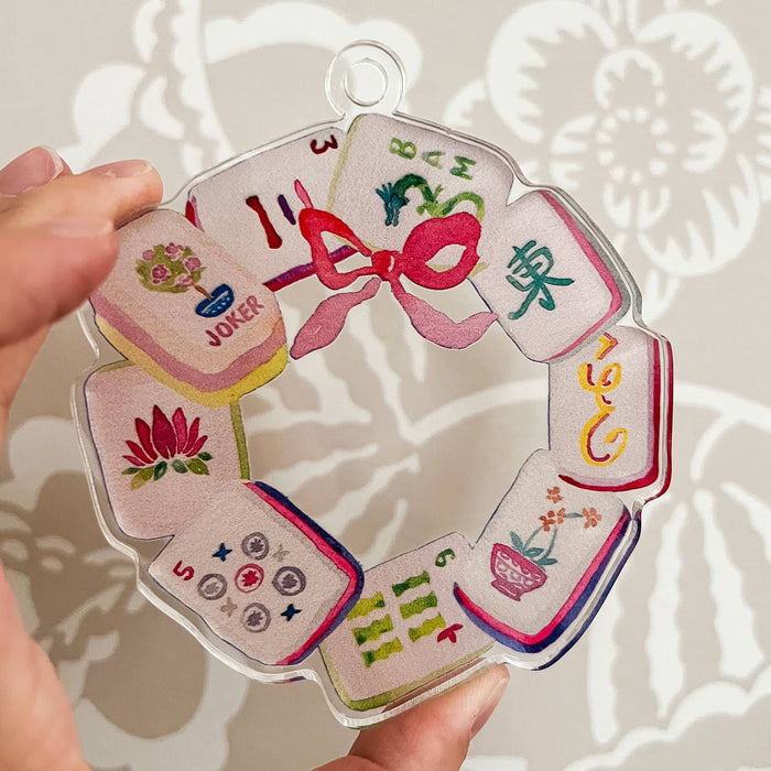 A hand holds a clear acrylic holiday ornament featuring colorful illustrations of mahjong tiles and a pink bow, set against a light patterned wall.