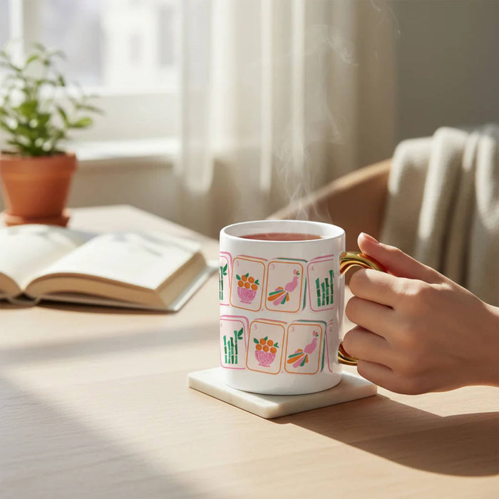 Person holding a mug with Mahjong tile illustrations on a table with a book and plant in the background.