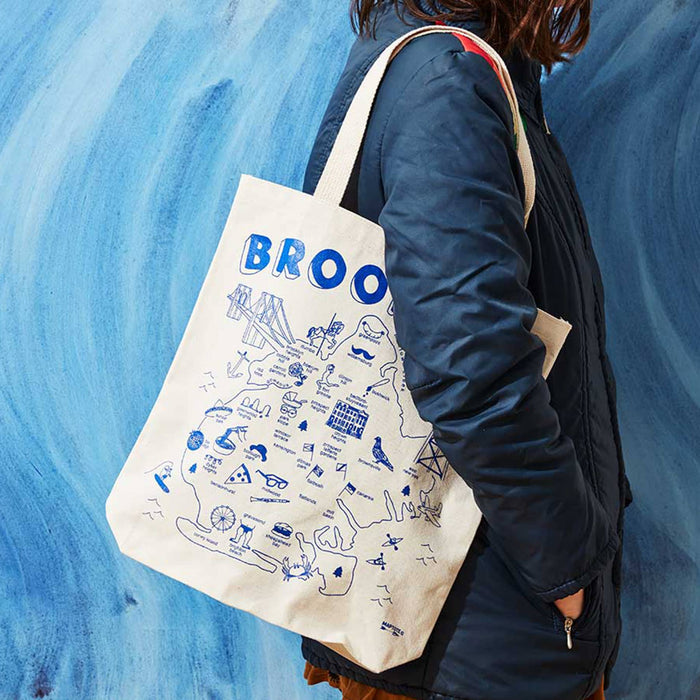 Person holding a cream tote bag with Brooklyn landmarks printed on it in blue, against a blue textured background.