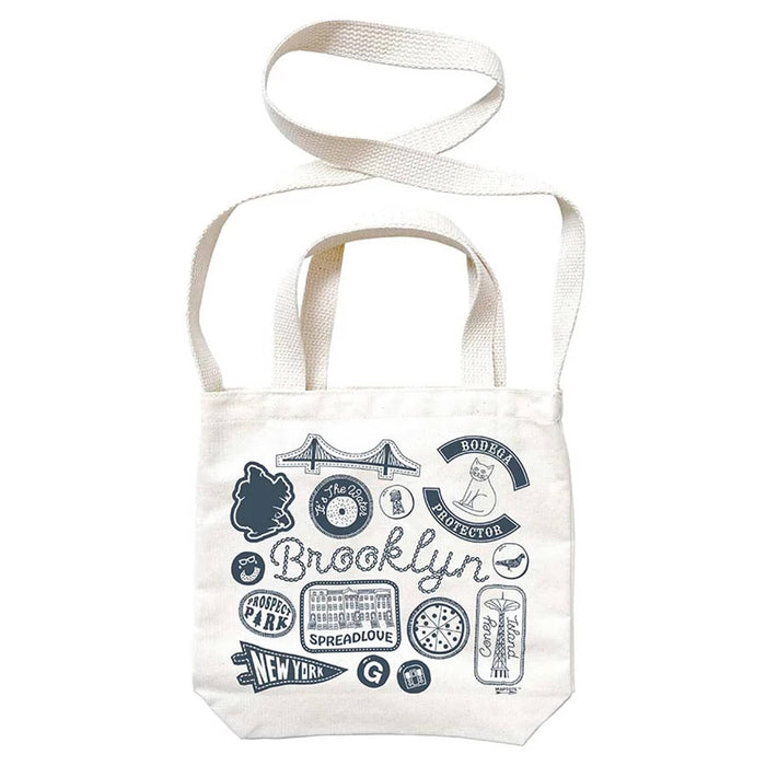 White tote bag with blue Brooklyn graphics and text, with white webbed handles and shoulder strap, on a white background.