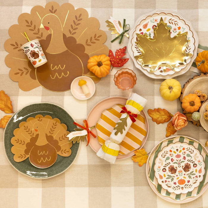 An autumn-themed table setting featuring ceramic and paper plates with turkey and maple leaf designs, a matching napkin holder, and fall decorations on a beige and white checkered tablecloth.