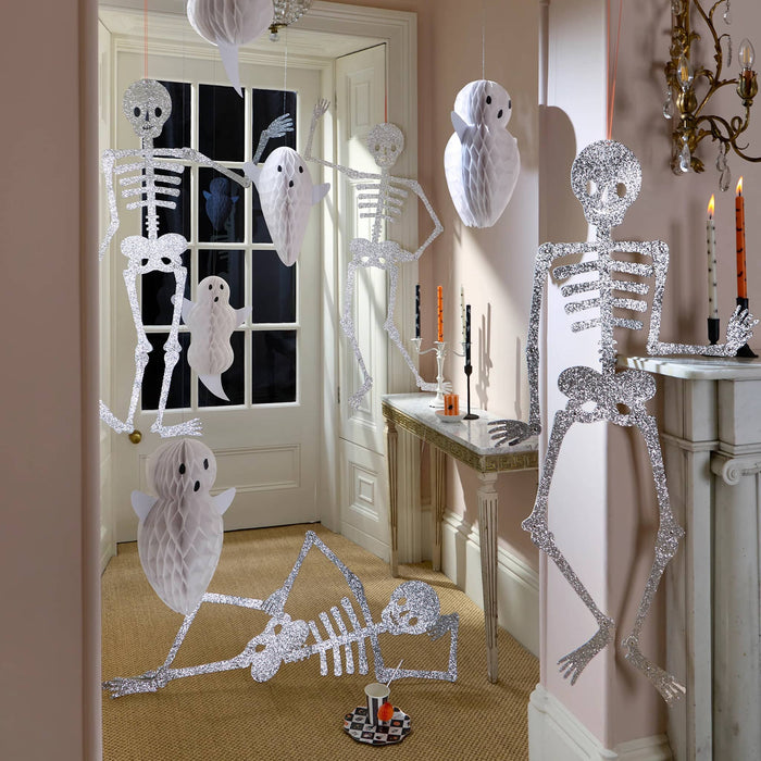 Halloween decorations featuring giant silver glitter skeleton figures, white ghost cutouts, and hanging white owl cutouts displayed in a decorated hallway with a fireplace and table.