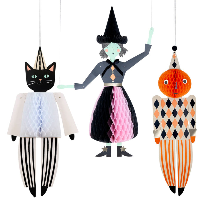 Three hanging decorative paper on a white background: a black cat in a pointed hat, a witch with a black hat and pink-and-black dress, and a pumpkin character with orange, black, and cream harlequin pattern.