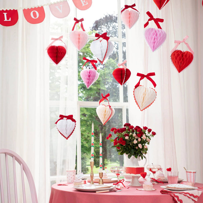 Meri Meri Honeycomb Hearts Valentine's Day decorations, 3 colors of hearts with velvet bows on top, hanging from gold thread over a table set for a party.