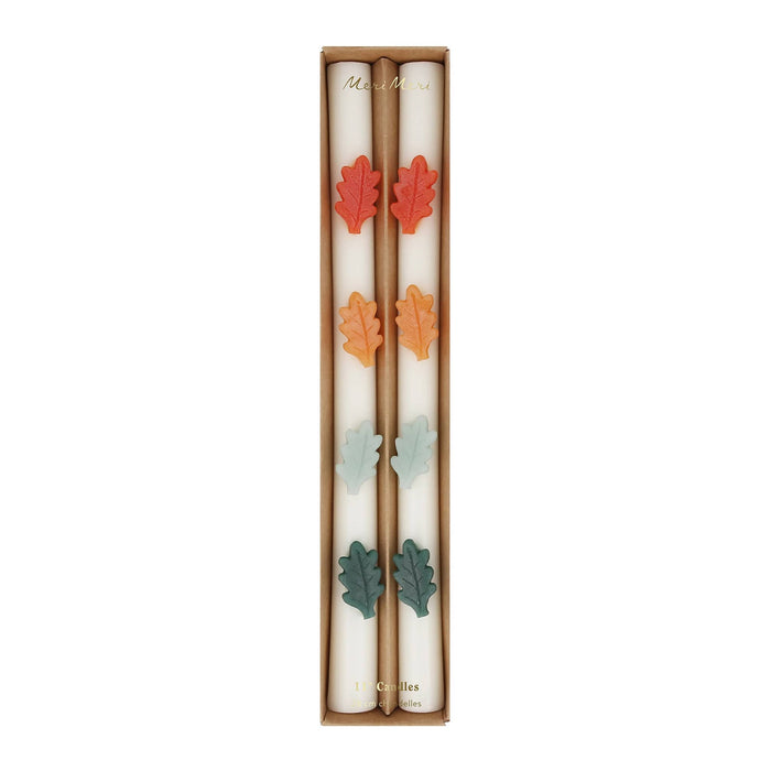 A boxed set of two cream-colored candles with decorative wax autumn leaves in red, orange, yellow, mint, and green, presented in a brown cardboard package on a white background.