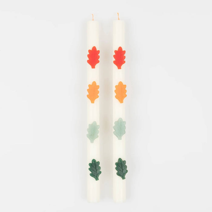 A boxed set of two cream-colored candles with decorative wax autumn leaves in red, orange, yellow, mint, and green, set against a white background.