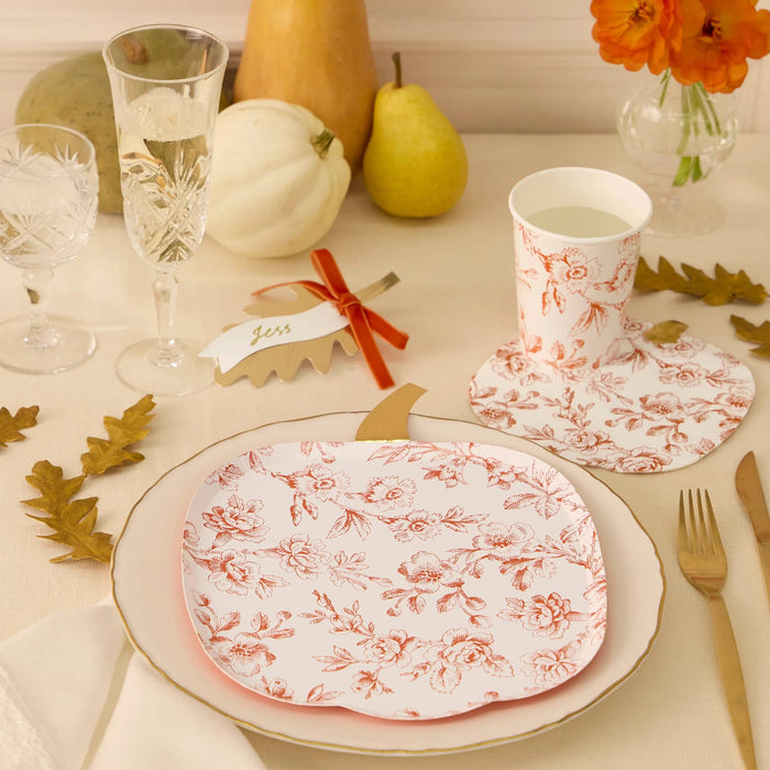 A styled tabletop setting featuring a pumpkin-shaped plate and napkin and a paper cup all with an orange floral toile pattern, arranged with gold-toned cutlery, glasses, fruit, and autumn leaves on a light cream surface.