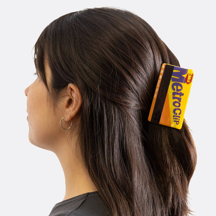 A woman shown in profile wearing a yellow and black MetroClip hair claw clip on her dark brown hair, against a light gray background.