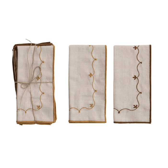 Three folded cream cotton napkins with brown and mustard embroidered floral pattern, shown on a plain white background. One napkin is tied with twine on the left, and two are shown.