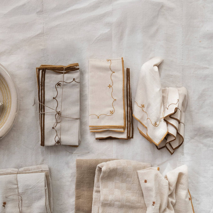 A styled flat-lay of multiple folded cotton napkins in cream, beige, and light brown, decorated with delicate embroidered floral vines and tied with twine on a textured beige background.
