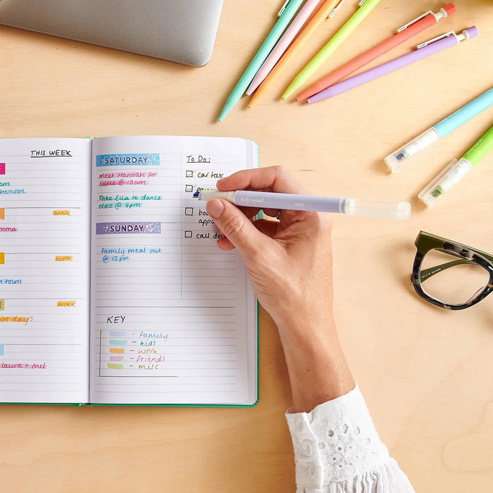 A top-down view of a person's hand writing in an open bullet journal with a purple pen and highlighter, surrounded by colorful pens, highlighters, and eyeglasses on a light wood desk.