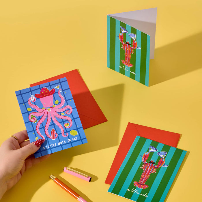 A hand holds a colorful illustrated card featuring a pink octopus on a blue grid background, with red envelopes and two more cards arranged on a bright yellow surface. The cards have playful lobster illustrations and striped green-and-blue backgrounds.