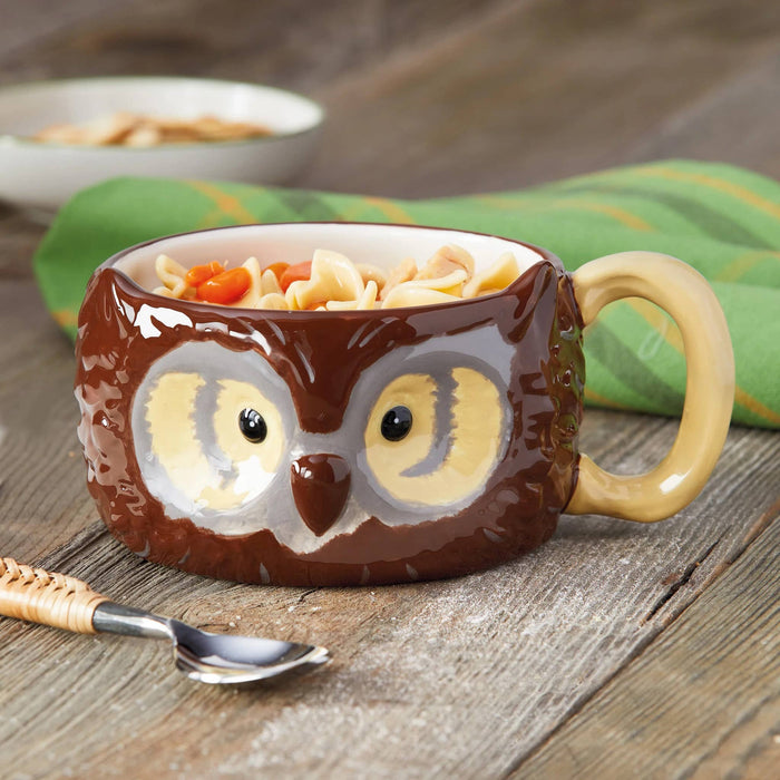 An owl-shaped ceramic mug filled with noodle soup, placed on a rustic wooden table with a spoon in the foreground and a green cloth in the background.