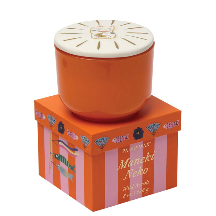 Paddywax Maneki Neko candle in an orange ceramic pot with a cream-colored lid, displayed on top of its orange and pink striped packaging box.