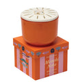 Paddywax Maneki Neko candle in an orange ceramic pot with a cream-colored lid, displayed on top of its orange and pink striped packaging box.
