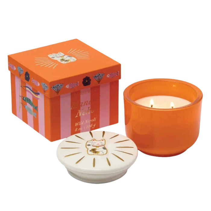 A lit orange scented candle in a translucent orange glass jar, shown with its matching orange box and a white decorative lid featuring a cat illustration, on a plain white background.