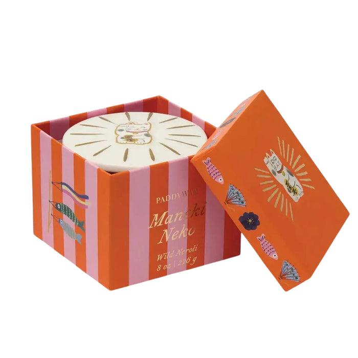 A round white soap bar with a gold cat illustration sits inside an open decorative box with pink and orange vertical stripes. The lid leans against the box, showing orange panels with additional cat and diamond illustrations.