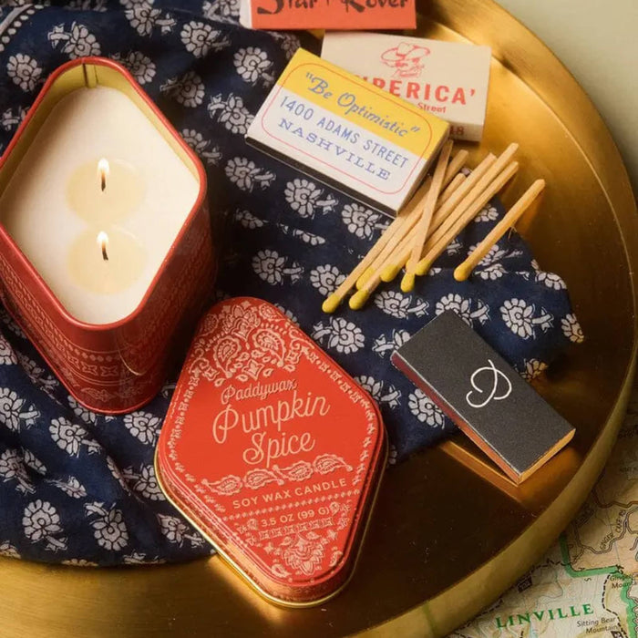 A styled flat-lay of a orange metal candle tin labeled 'Pumpkin Spice' beside a lit candle in a matching orange container, with vintage-style matches and a small black matchbox arranged on a gold tray over a navy floral cloth.