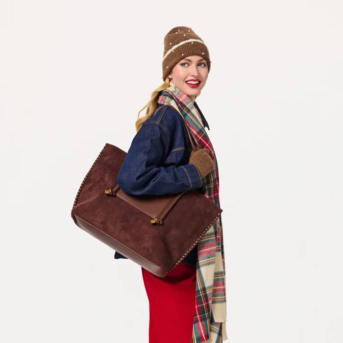 A woman wearing a brown knit beanie, blue denim jacket, red pants, and a green plaid scarf carries a large brown vegan suede tote bag with gold-tone hardware and a matching brown leather strap, set against a plain white background.