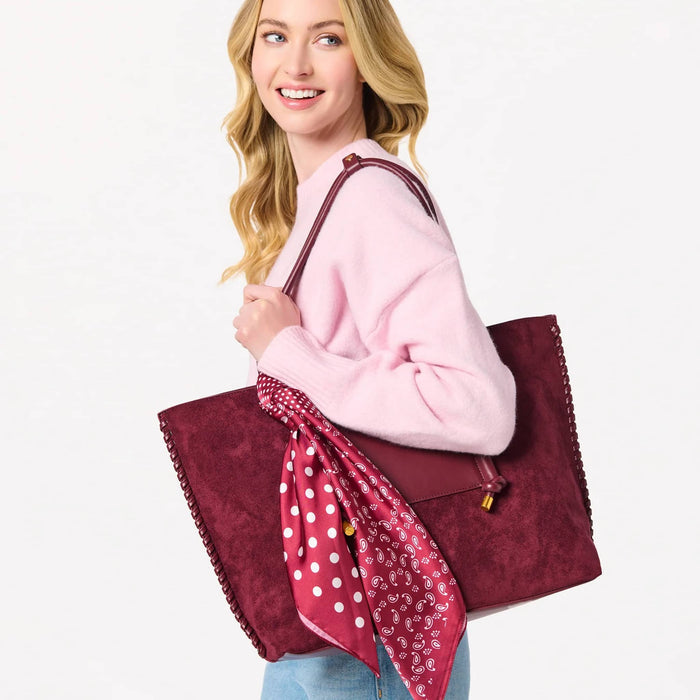 A woman wearing a pink sweater and blue jeans carries a large burgundy vegan suede tote bag with a matching shoulder strap. A pink polka-dot scarf is draped over the front of the bag.