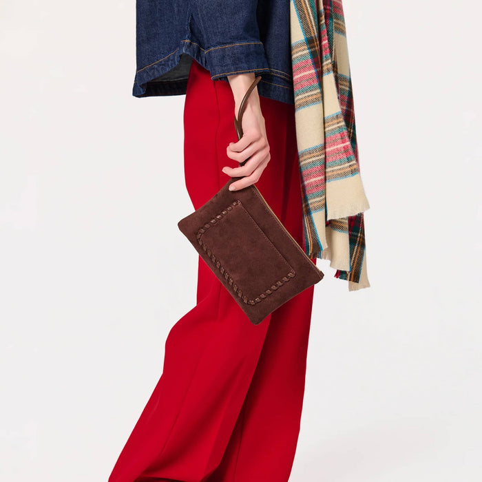 A person wearing bright red wide-leg pants, a dark blue denim jacket, and a multicolor plaid scarf, holding a brown vegan suede clutch bag with a matching wristlet strap against a plain white background.