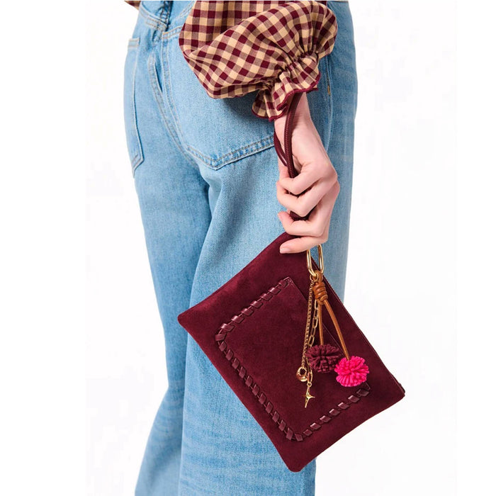 A person wearing light blue jeans holds a burgundy vegan suede clutch bag with a brown vegan leather strap, decorative stitching, gold-tone charms, and a bright pink pom-pom accent against a plain white background.