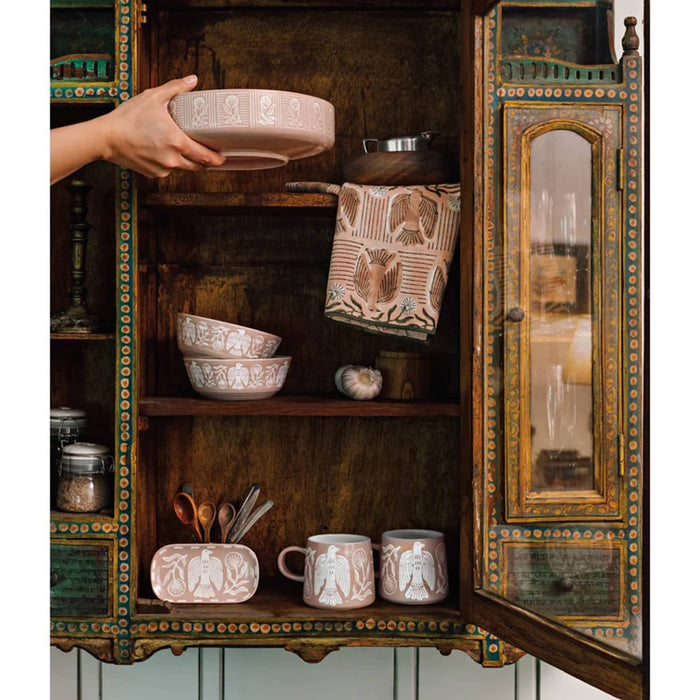 Hand holding a pink ceramic bowl with embossed design in front of an ornate wooden cabinet with other items from the Danica Peregrine collection.