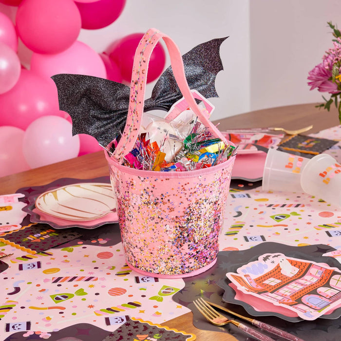 A pink glittery fabric bucket filled with candy and small black bat wing bow, styled on a decorated party table with pink balloons, paper plates, and a flower vase.