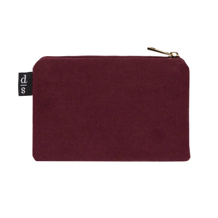 Back of a rectangular burgundy canvas zippered pouch with a small black fabric tag on the left side featuring a white 'ds' logo, set against a plain white background.