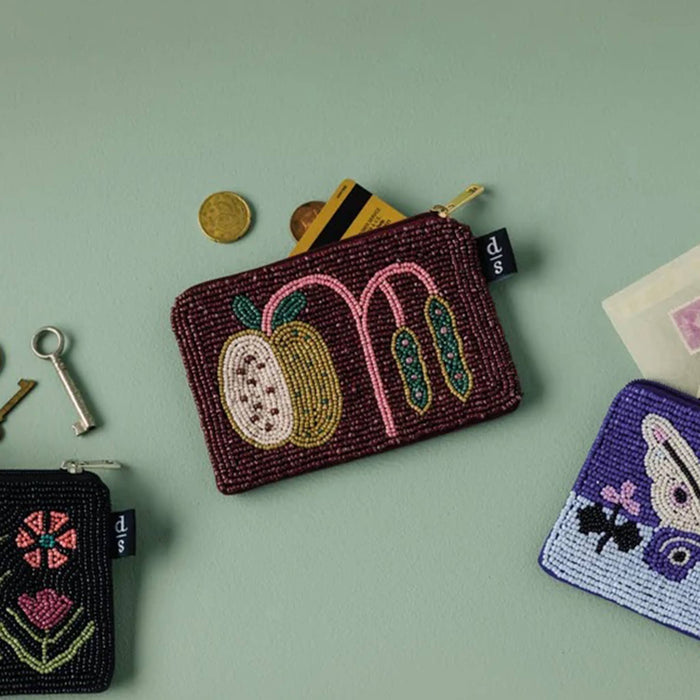 A hand-beaded coin purse in burgundy with embroidered fruit and leaf motifs, styled on a sage green background with keys, coins, cards, and other purses.