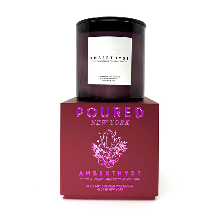 A dark amber glass candle jar with a white label reading AMBERTHYST, displayed on top of a deep red rectangular box with pink foil branding text and crystal illustration. The box reads POURED NEW YORK and AMBERTHYST.