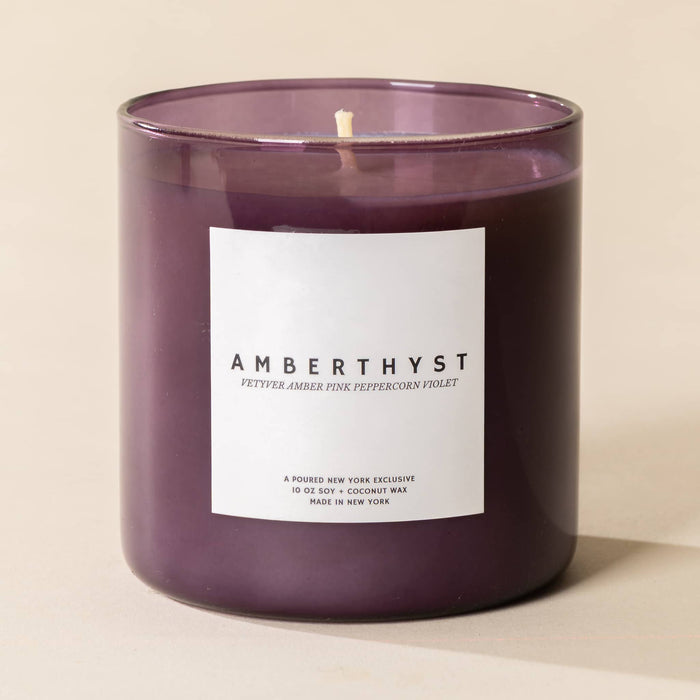 A purple glass jar candle with a white rectangular label reading 'AMBERTHYST' and smaller scent note text, photographed on a warm beige background.