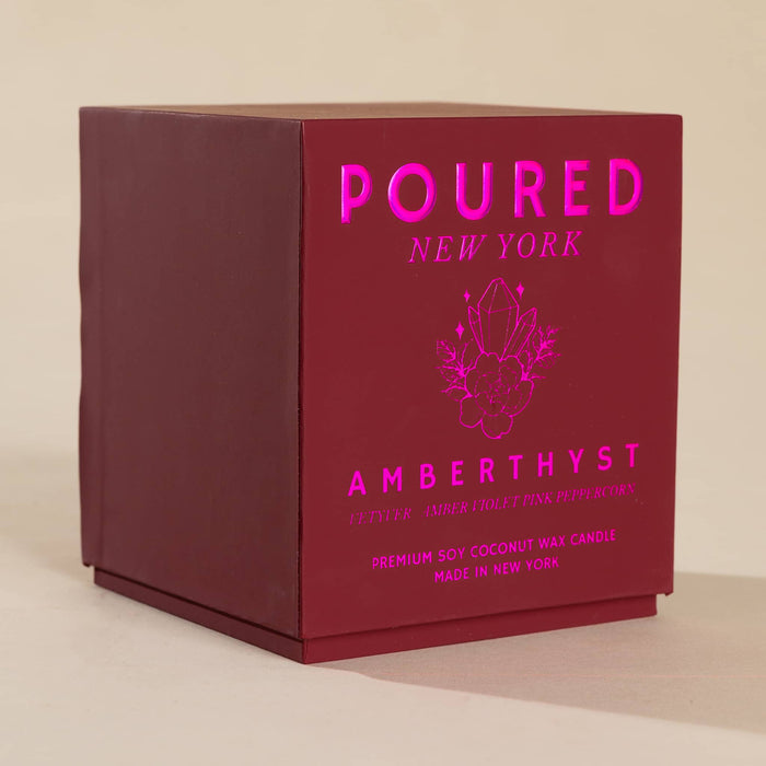 A dark red square product box for a Poured New York Amberthyst candle, featuring pink foil text and a crystal graphic on a beige background.