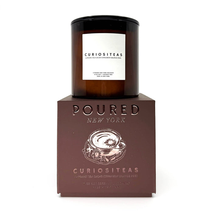 An amber glass jar candle with a black lid and white label reading 'CURIOSITEAS' sits on top of a dark brown embossed box labeled 'POURED NEW YORK' and 'CURIOSITEAS'.