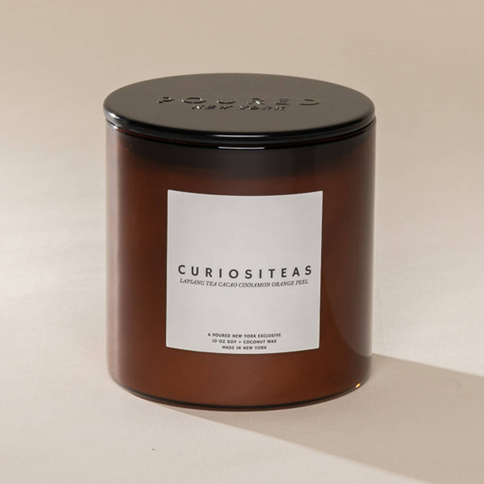 An amber glass jar candle with a black metal lid and a white label reading 'CURIOSITEAS' on a warm beige background.